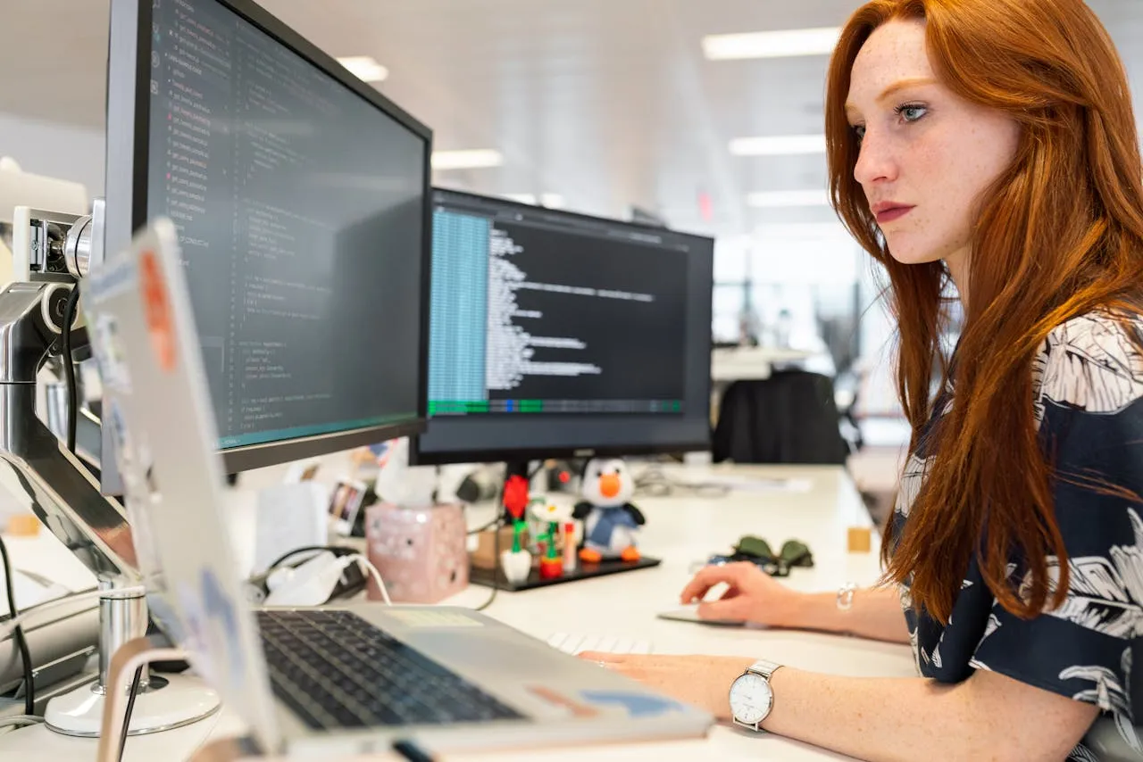 Woman working at a laptop with two monitors showing code in an open office