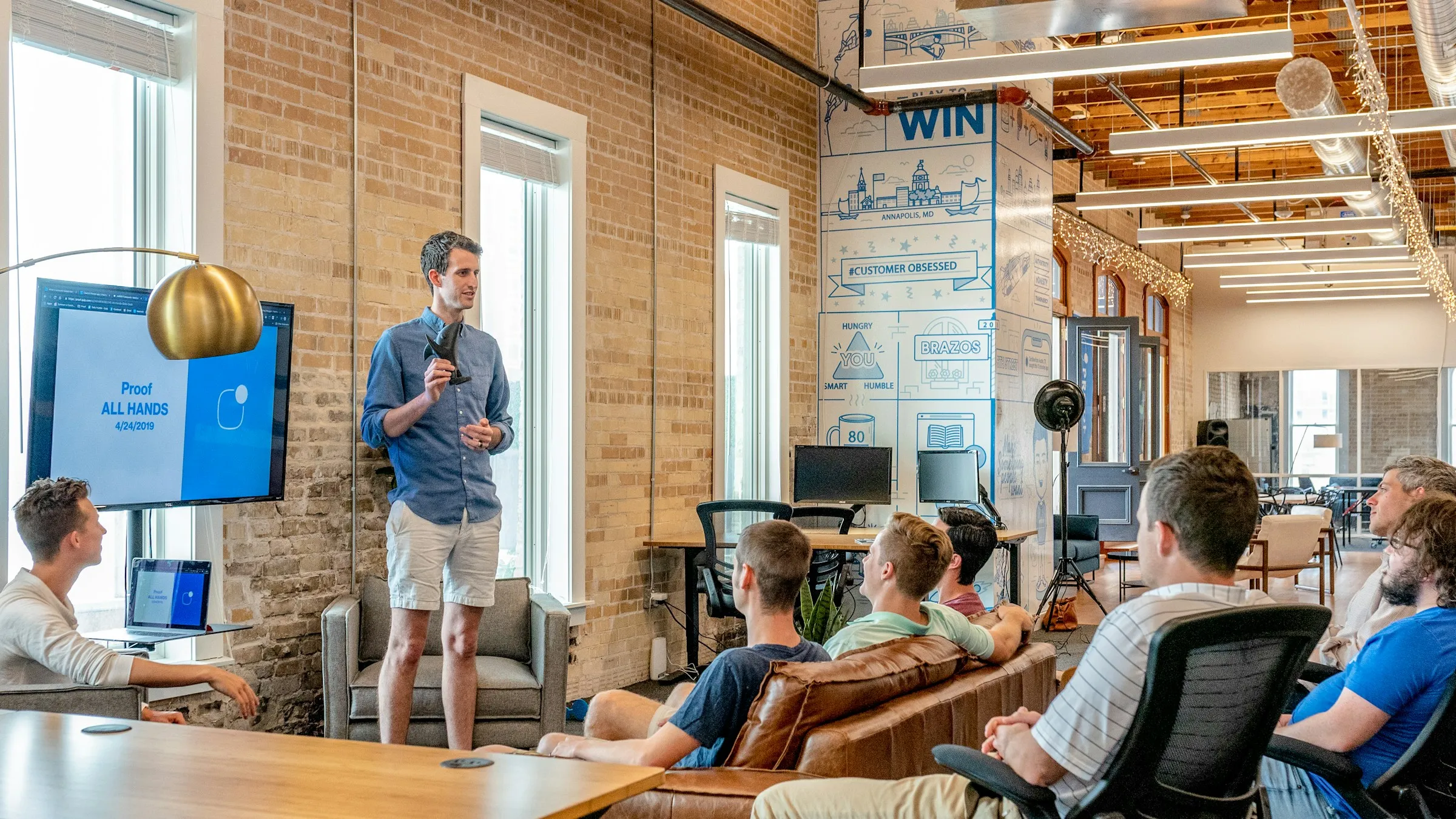 A startup team gathered on sofas in a brick-walled office while a colleague presents