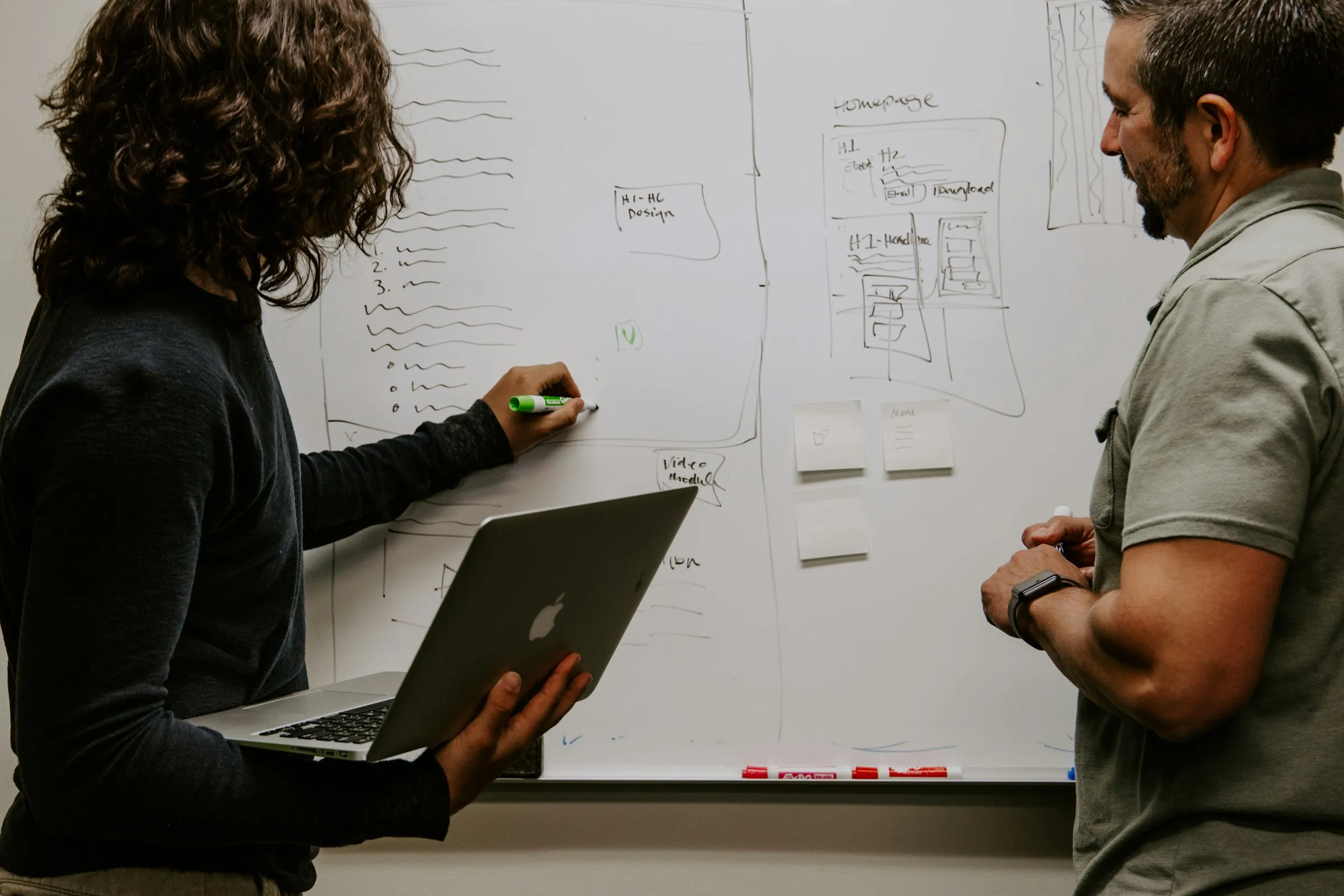 Two people reviewing wireframes sketched on a whiteboard, one holding a laptop