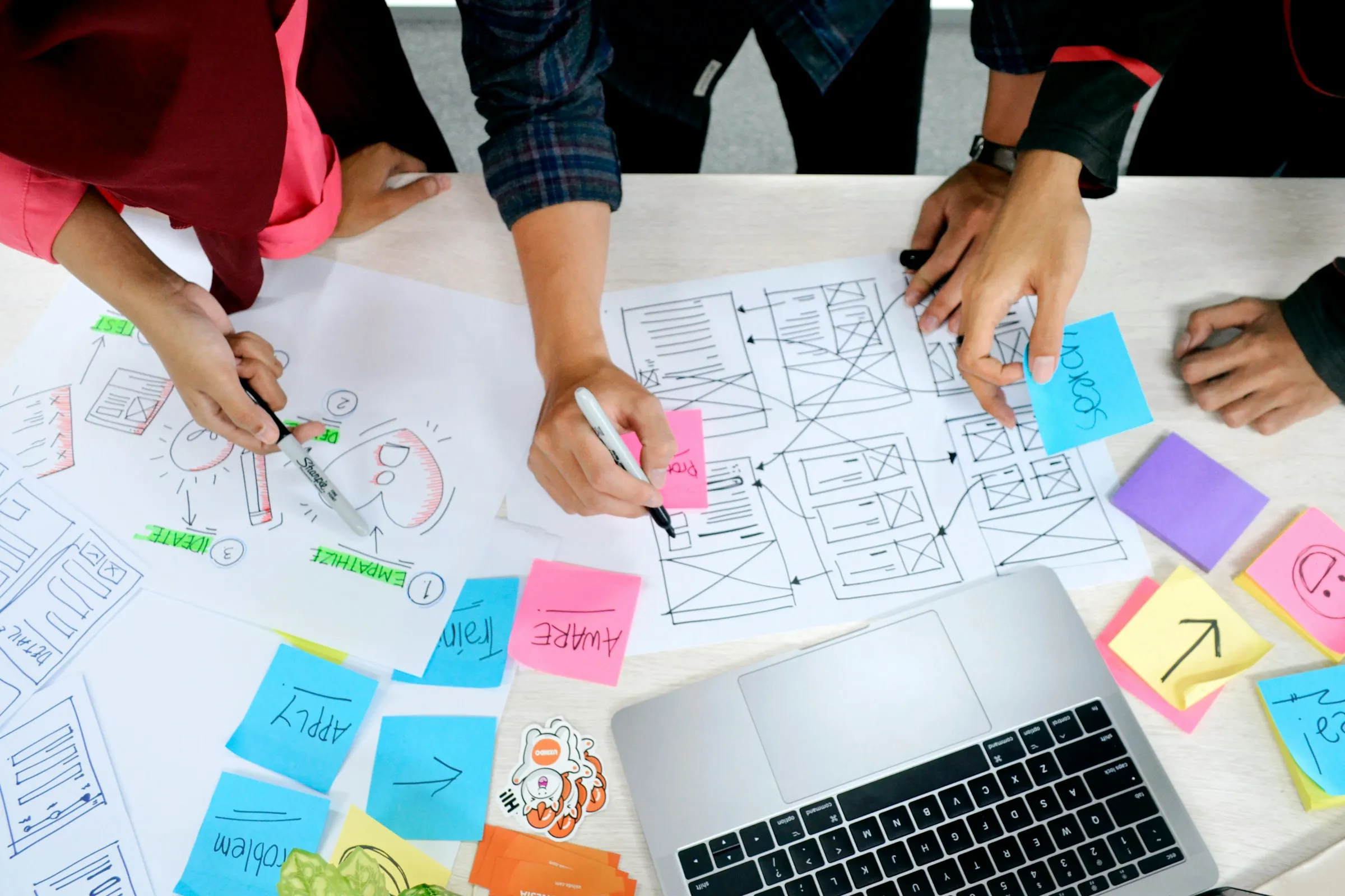 Overhead view of a team sketching wireframes on paper with sticky notes and a laptop on the table